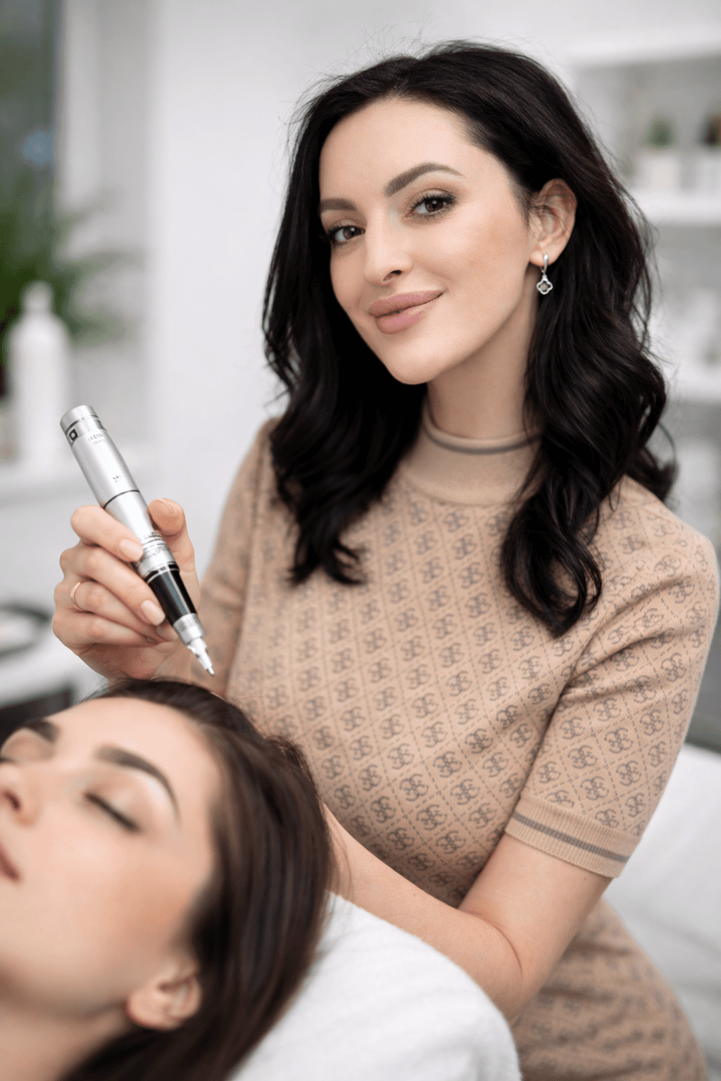 Brunette beautician holding a silver permanent makeup pen over a client's face in a studio.
